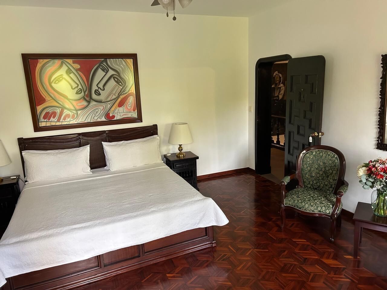 Une chambre d’hôtel avec un grand lit, une table de chevet, une chaise, un tableau au mur et une porte donnant sur une autre pièce.