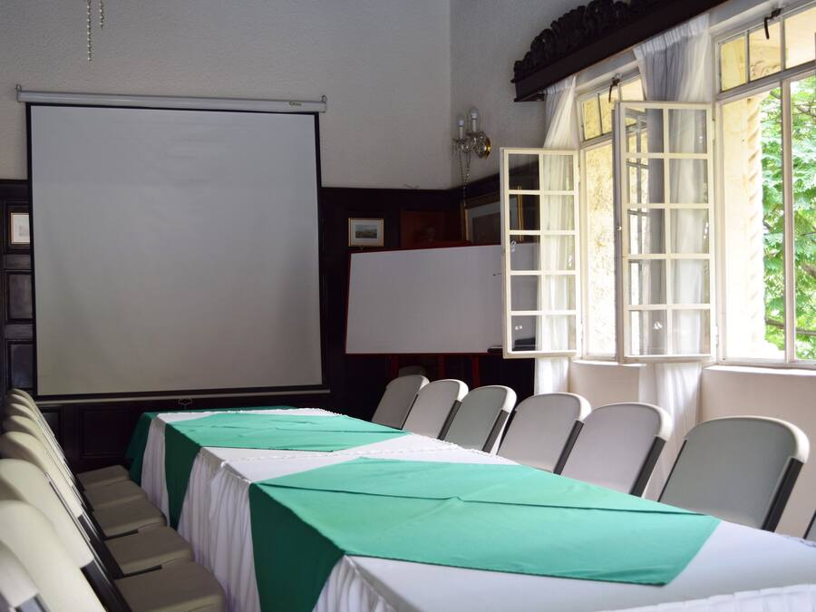 Una sala de conferencias con una mesa larga, sillas, una pantalla de proyector y ventanas con cortinas.