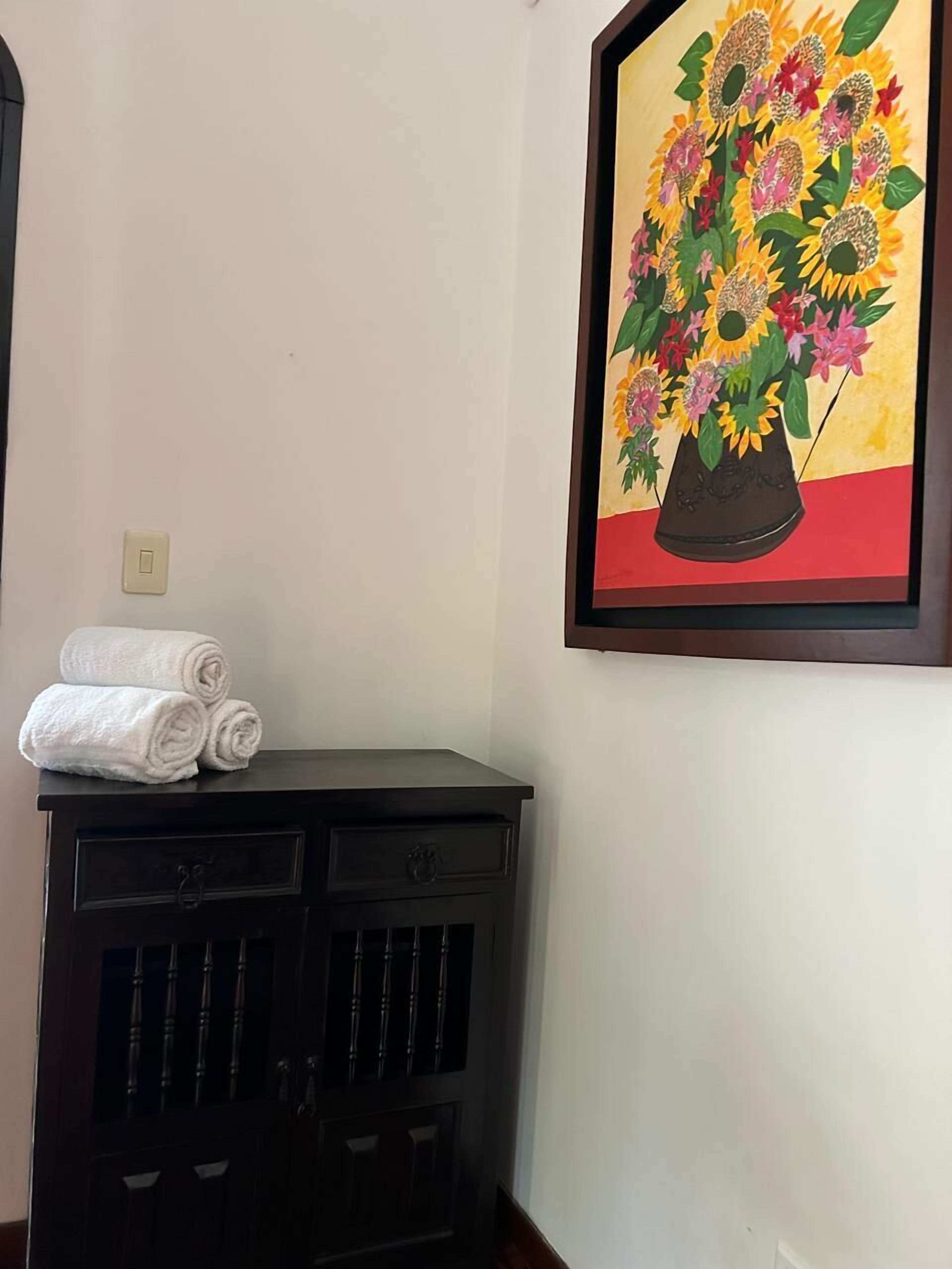 Une commode en bois foncé, et au-dessus, accrochée au mur, une peinture encadrée représentant une composition florale.