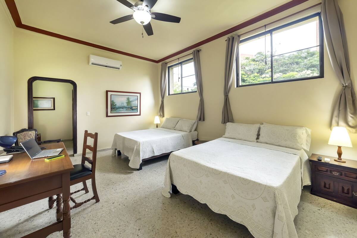 A bedroom with two beds, a desk with a laptop, a chair, and a nightstand with a lamp.
