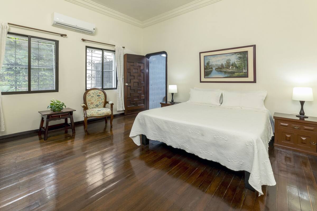 A bedroom with a large bed, wooden flooring, two windows with curtains, a chair, a bedside table with a lamp, and a painting on the wall.