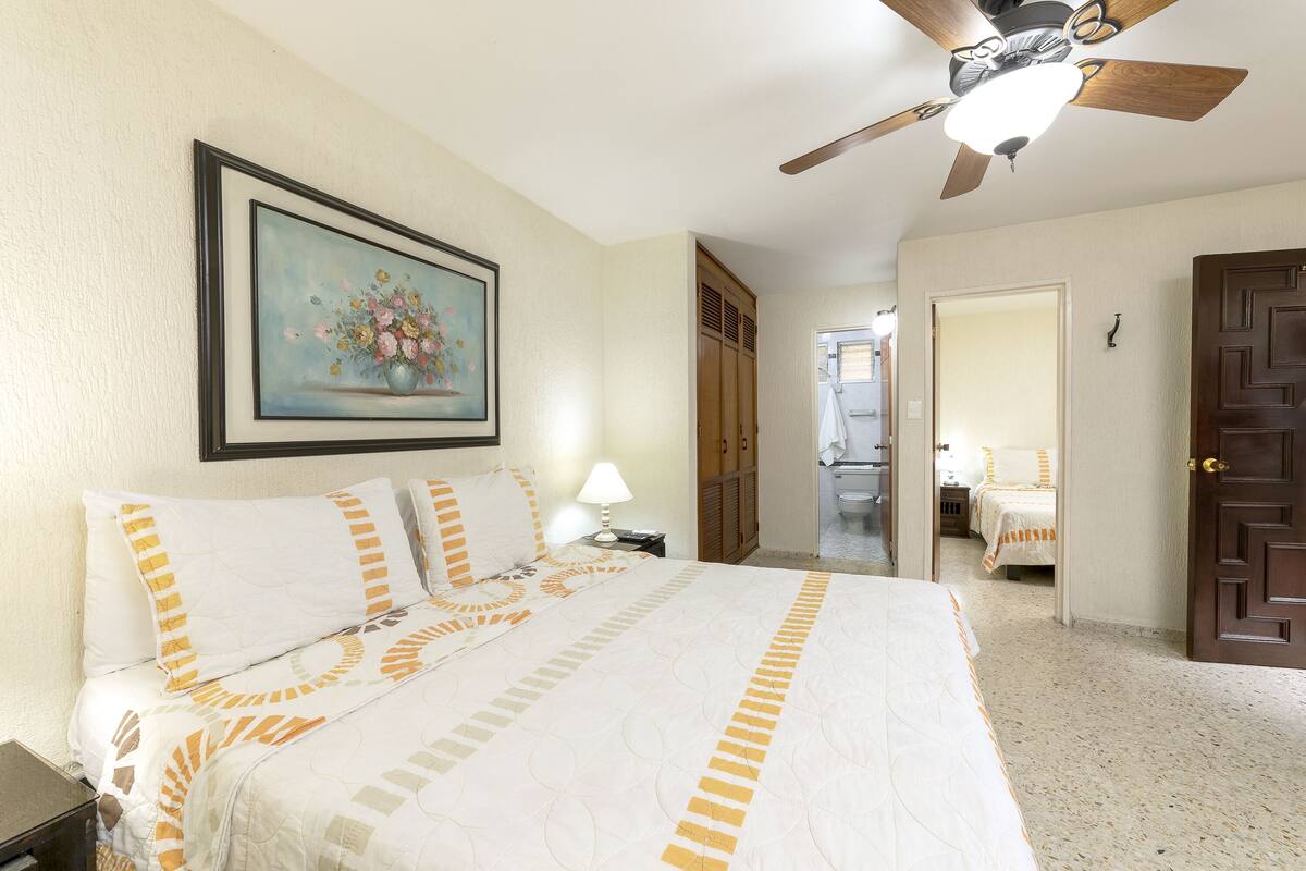 A hotel room with a bed, a ceiling fan, a painting, and a door leading to another room.