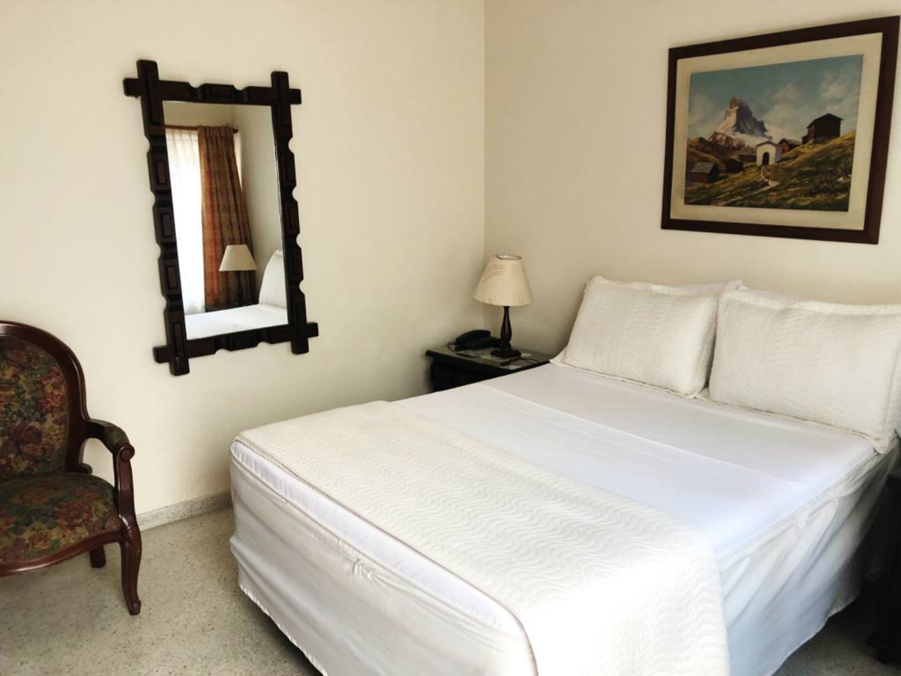 Una cama bien hecha con ropa de cama blanca, una mesita de noche de madera con una lámpara, una silla con estampado floral, un cuadro enmarco de un paisaje de montaña y un espejo con marco oscuro.