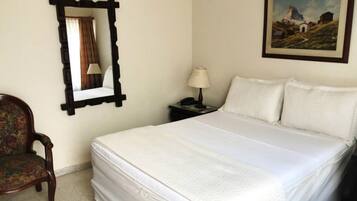 Una cama bien hecha con ropa de cama blanca, una mesita de noche de madera con una lámpara, una silla con estampado floral, un cuadro enmarco de un paisaje de montaña y un espejo con marco oscuro.