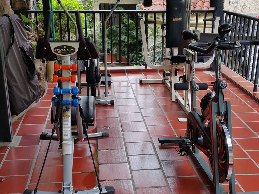 Ein überdachter Outdoor-Fitnessbereich mit verschiedenen Trainingsgeräten, darunter Laufband, Crosstrainer und Kraftgeräte.
