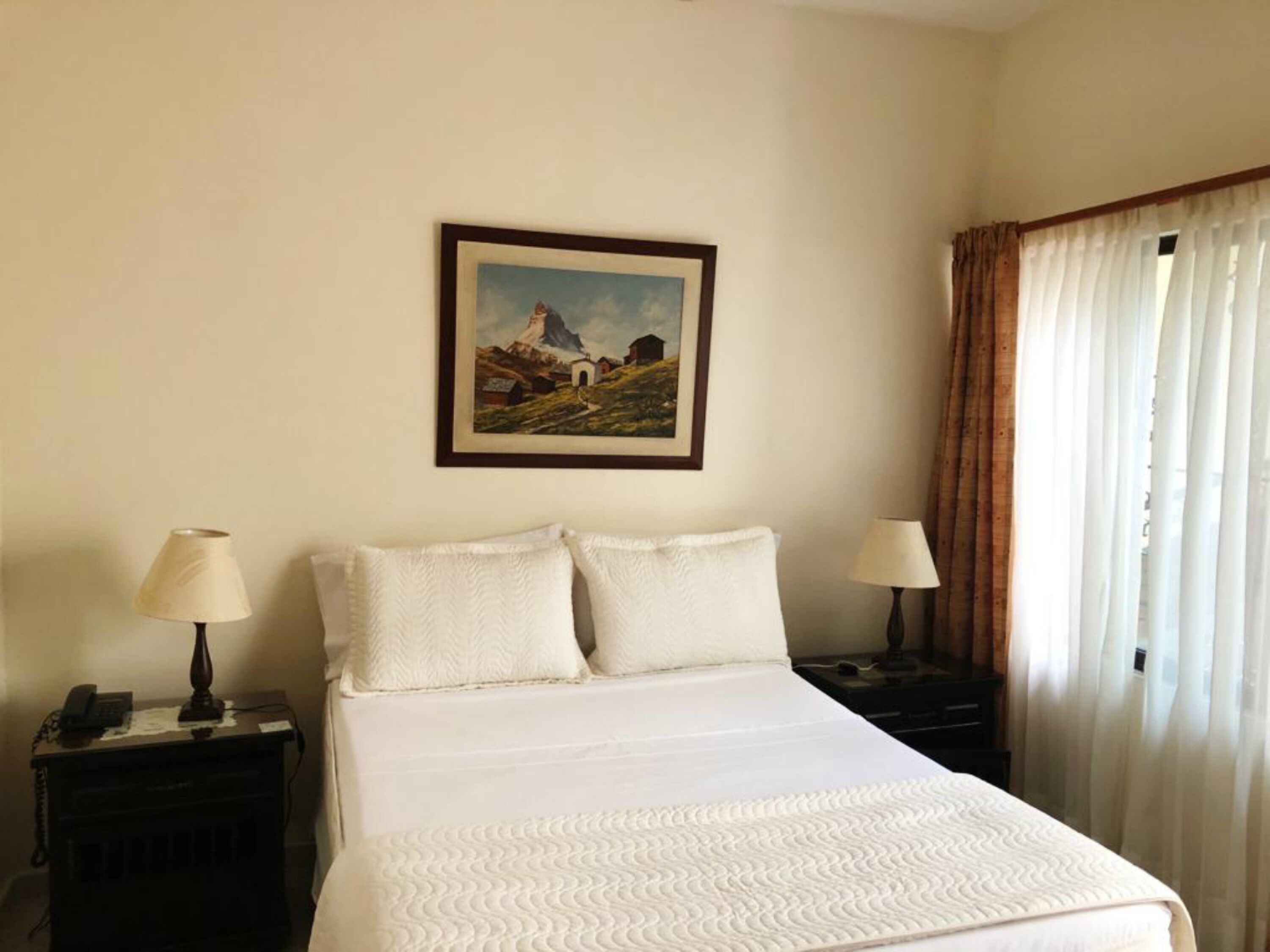 Habitación de hotel con una cama, dos mesitas de noche con lámparas, un cuadro en la pared y una ventana con cortinas.