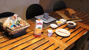 Uma mesa de madeira com uma cesta de lanches, um carton de suco de laranja, uma pequena tigela de frutas, um prato com um sanduíche e uma xícara.