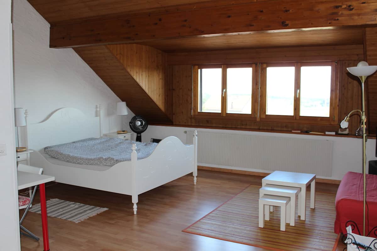 A bedroom with a sloped ceiling, a bed, a desk, a chair, a small table, and a window with a view.