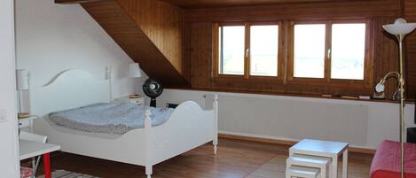 A bedroom with a sloped ceiling, a bed, a desk, a chair, a small table, and a window with a view.