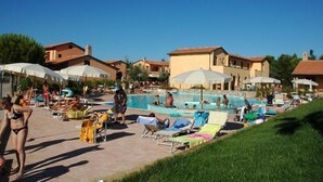 Zona de piscina con sillones y sombrillas, rodeada de edificios de estilo mediterráneo.