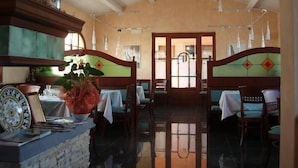 Un restaurante con techo alto, vigas de madera y un acceso central que da al exterior. Las mesas están dispuestas con manteles blancos, y sillas alrededor.