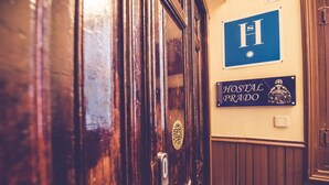 A wooden door with a keyhole and a brass handle, a sign with a large 'H' and a sun symbol, and a sign that reads "HOSTAL PRADO". - Hostal Prado (Madrid)