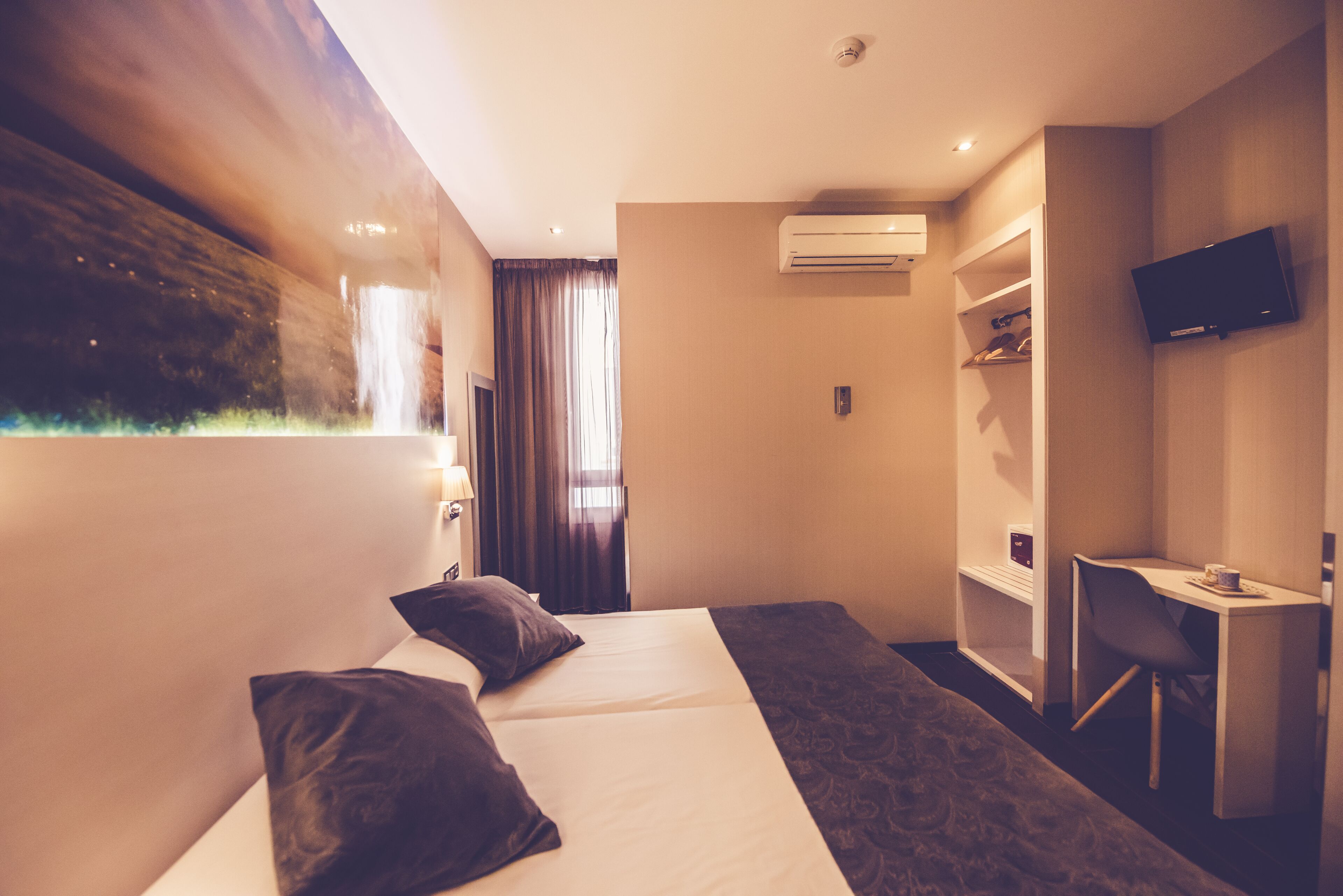 A hotel room with two beds, a desk, a TV, and an air conditioning unit.