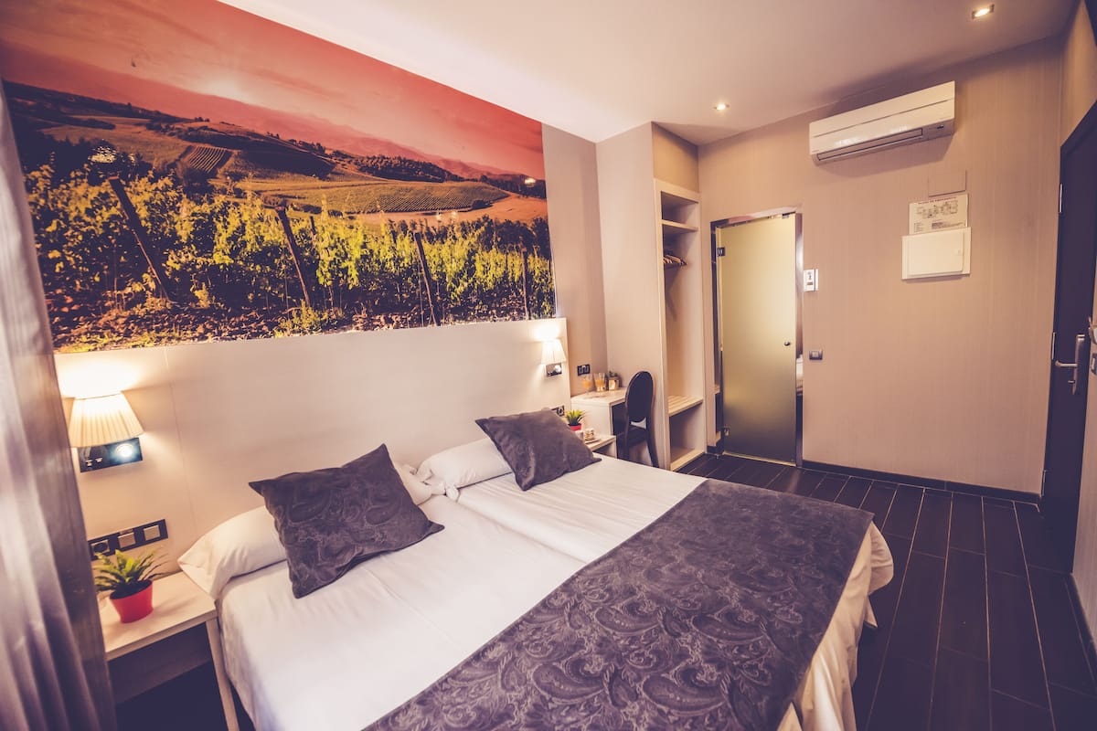 A hotel room with a large bed, a nightstand with a lamp, a potted plant, and a wall-mounted air conditioner.