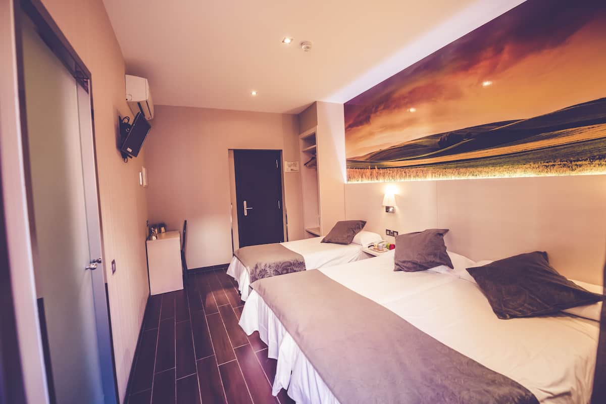 A hotel room with two beds, a painting on the wall, a door, and a small table with a lamp.