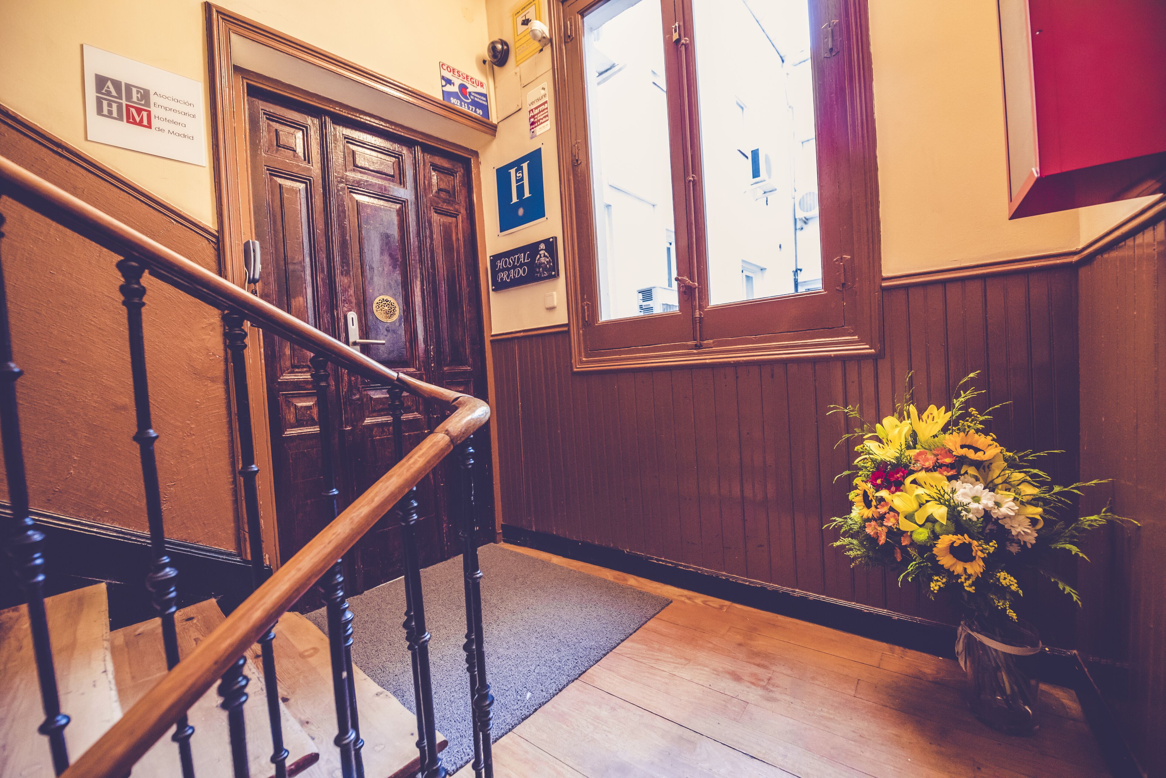 Una escalera de madera con barandilla, una puerta de madera y una ventana con vistas a edificios. Hay un cartel en la pared y un jarrón de flores en el suelo.