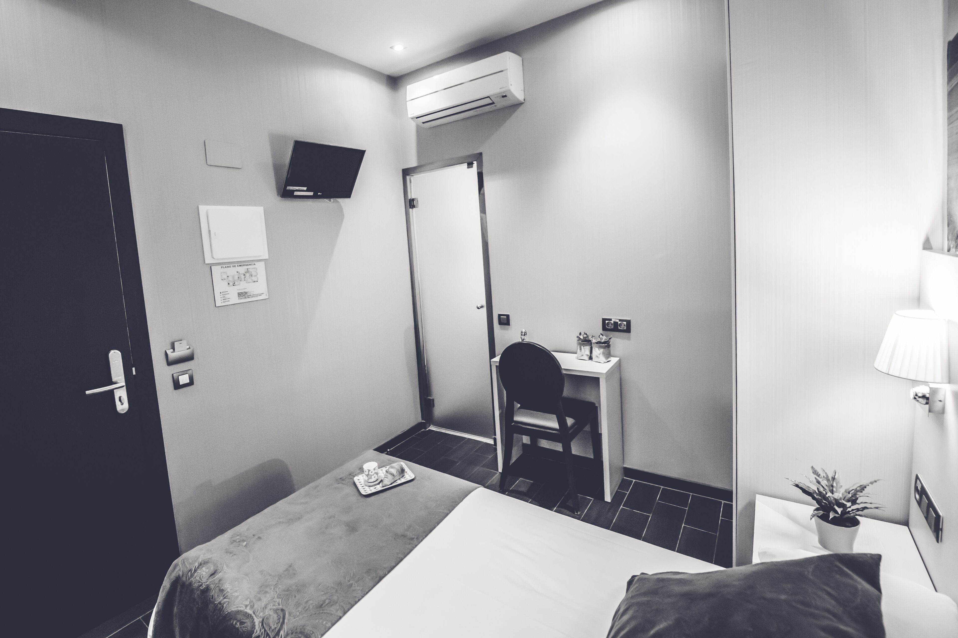 Une chambre d’hôtel comprenant un lit, un bureau, une chaise, une petite table avec un plateau de petit-déjeuner, un climatiseur, une télévision, une porte et une plante en pot.
