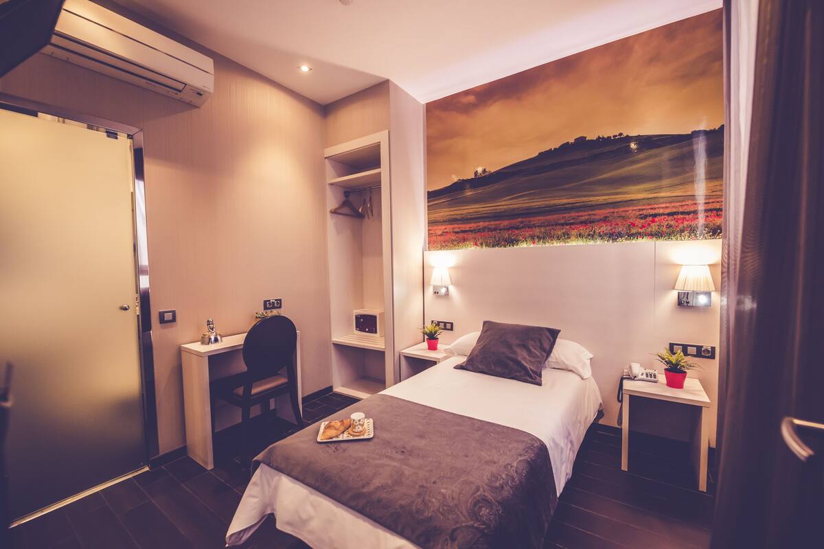 A hotel room with a bed, a desk, a chair, a nightstand, a lamp, and a wall mural.