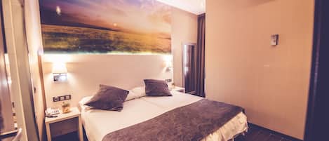 Una habitación de hotel con una cama grande, una mesita de noche con lámpara, aire acondicionado de pared y un mural de un paisaje.