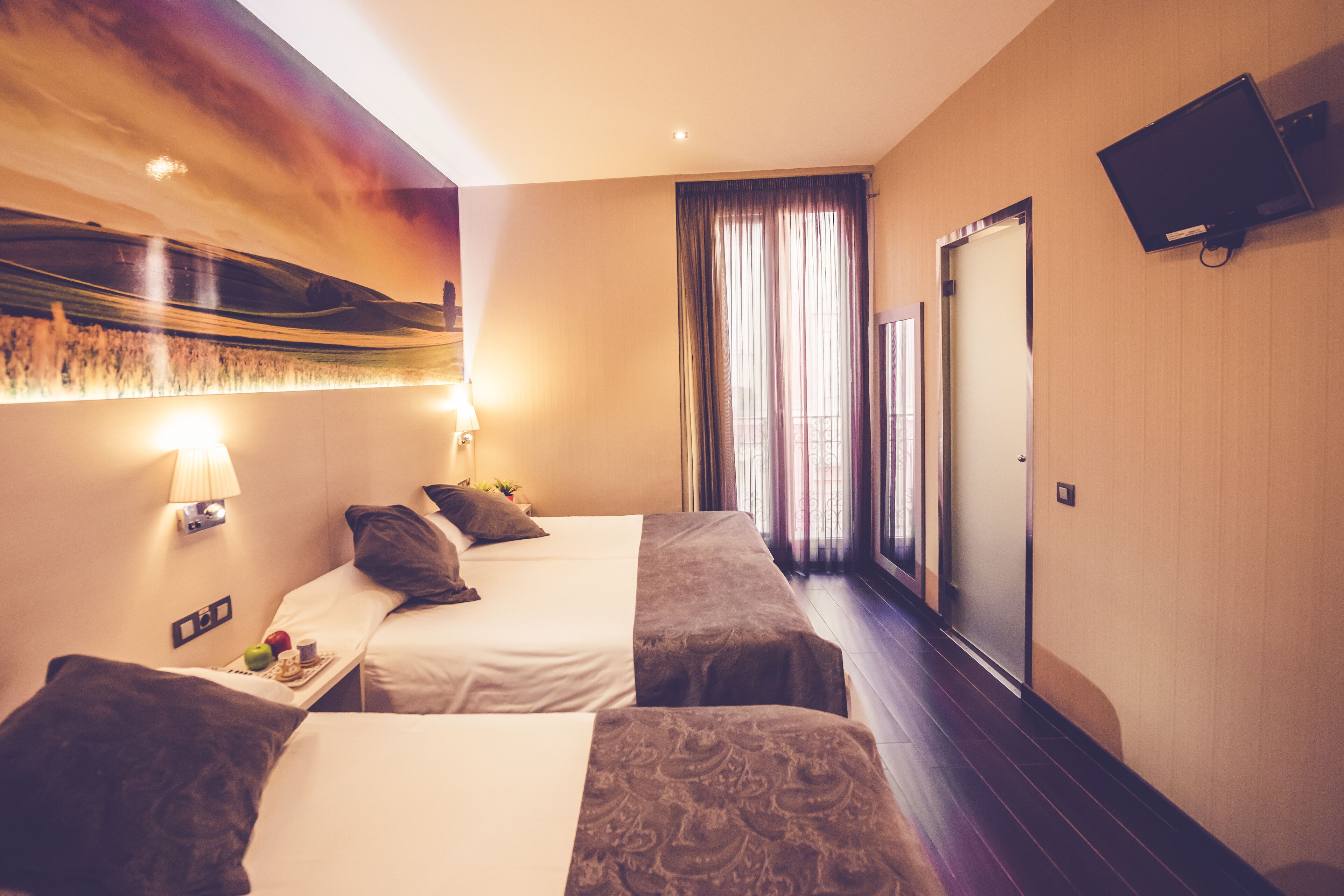 A hotel room with two beds, a flat-screen TV mounted on the wall, a window with curtains, and a wall-mounted lamp.