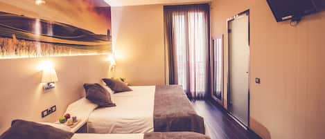 Habitación de hotel con dos camas, un televisor de pantalla plana montado en la pared, una ventana con cortinas y una lámpara de pared.