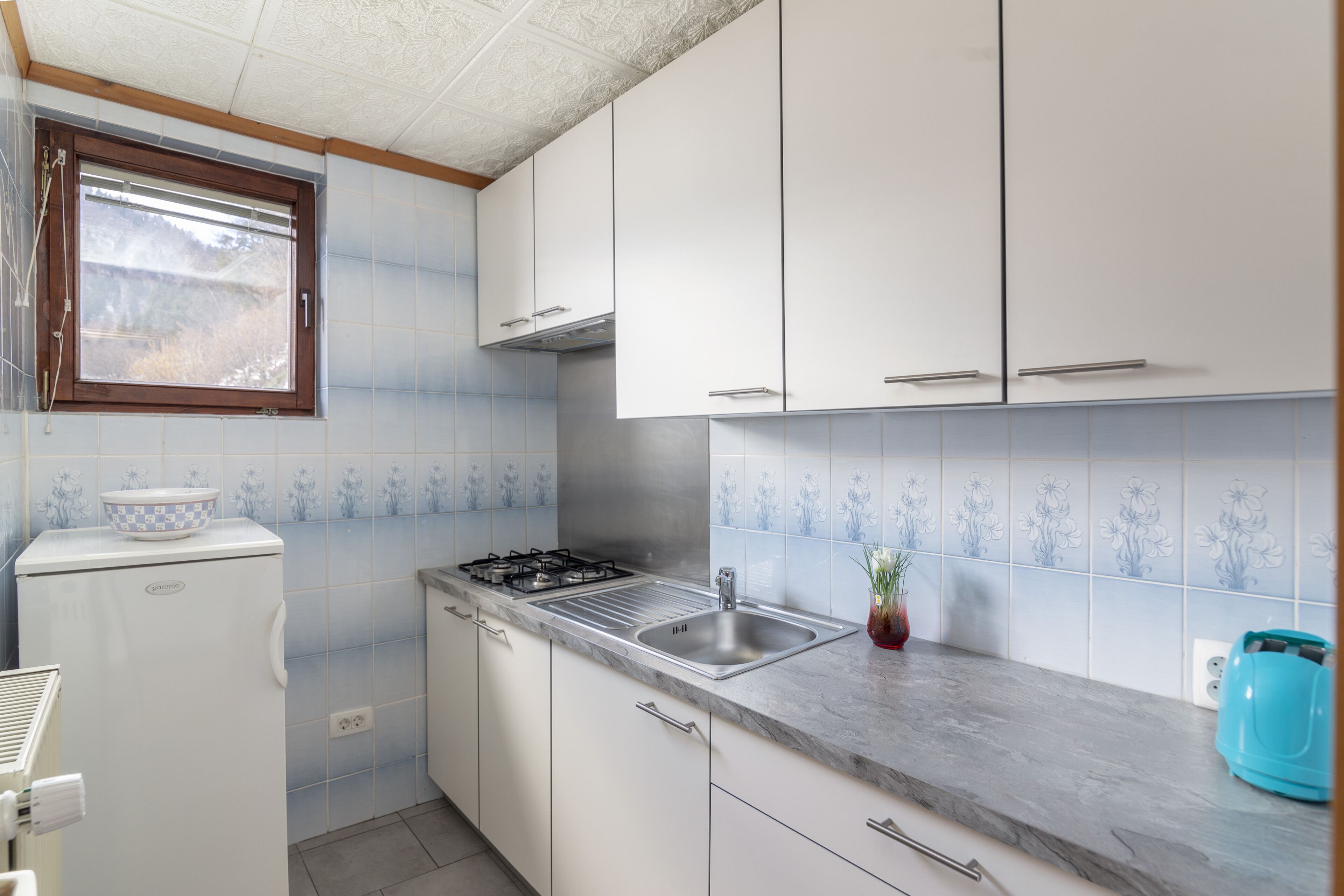 Cocina compacta con armarios blancos, estufa a gas empotrada, un lavamanos y una ventana con vistas al exterior.