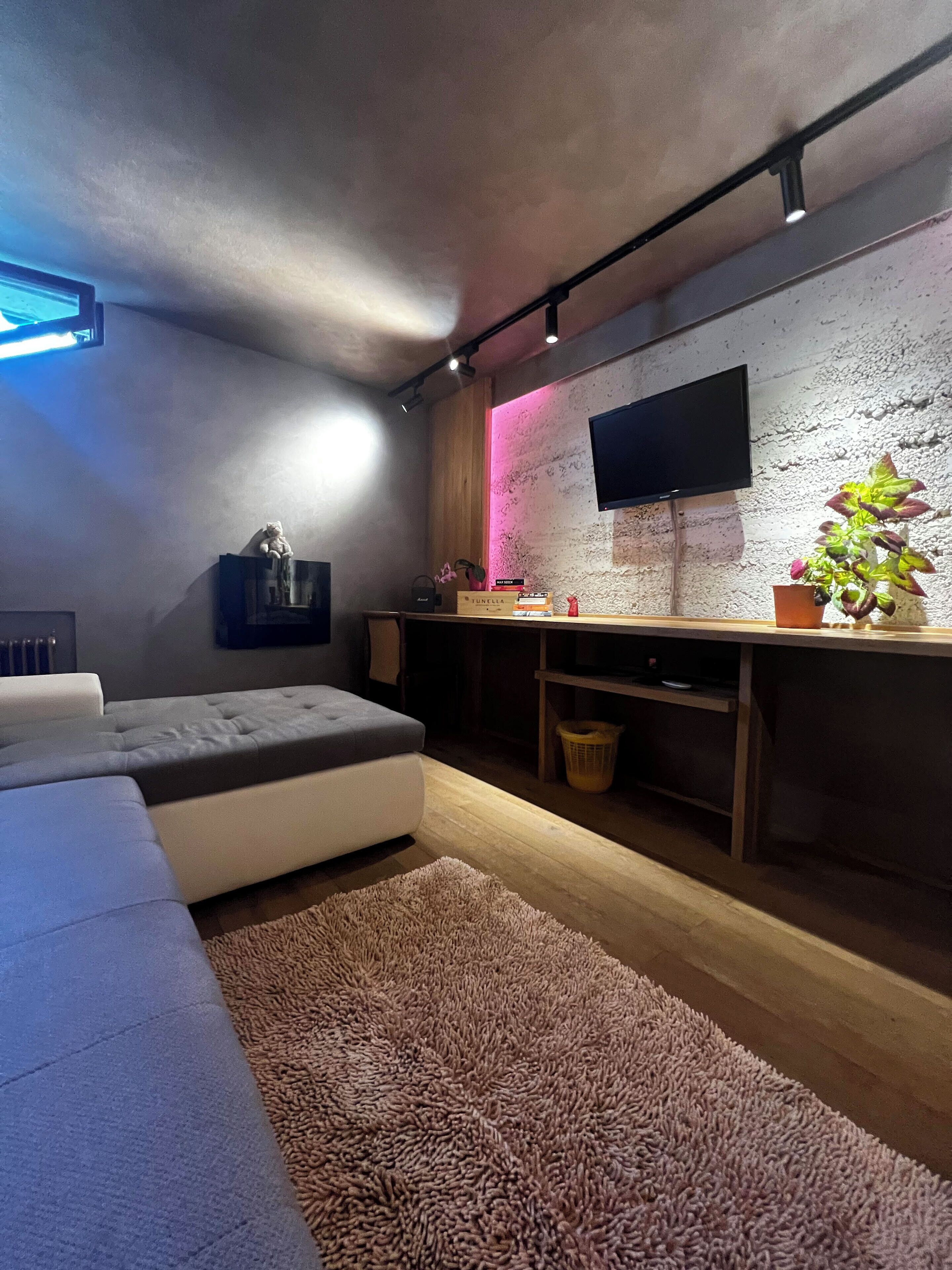 Ett modernt vardagsrum med en grå soffa, en platt-TV som är monterad på en vägg med en intressant yta, ett litet träbord med dekorativa föremål och en rosa ljuslist som ger en behaglig atmosfär.