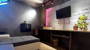 Ett modernt vardagsrum med en grå soffa, en platt-TV som är monterad på en vägg med en intressant yta, ett litet träbord med dekorativa föremål och en rosa ljuslist som ger en behaglig atmosfär.