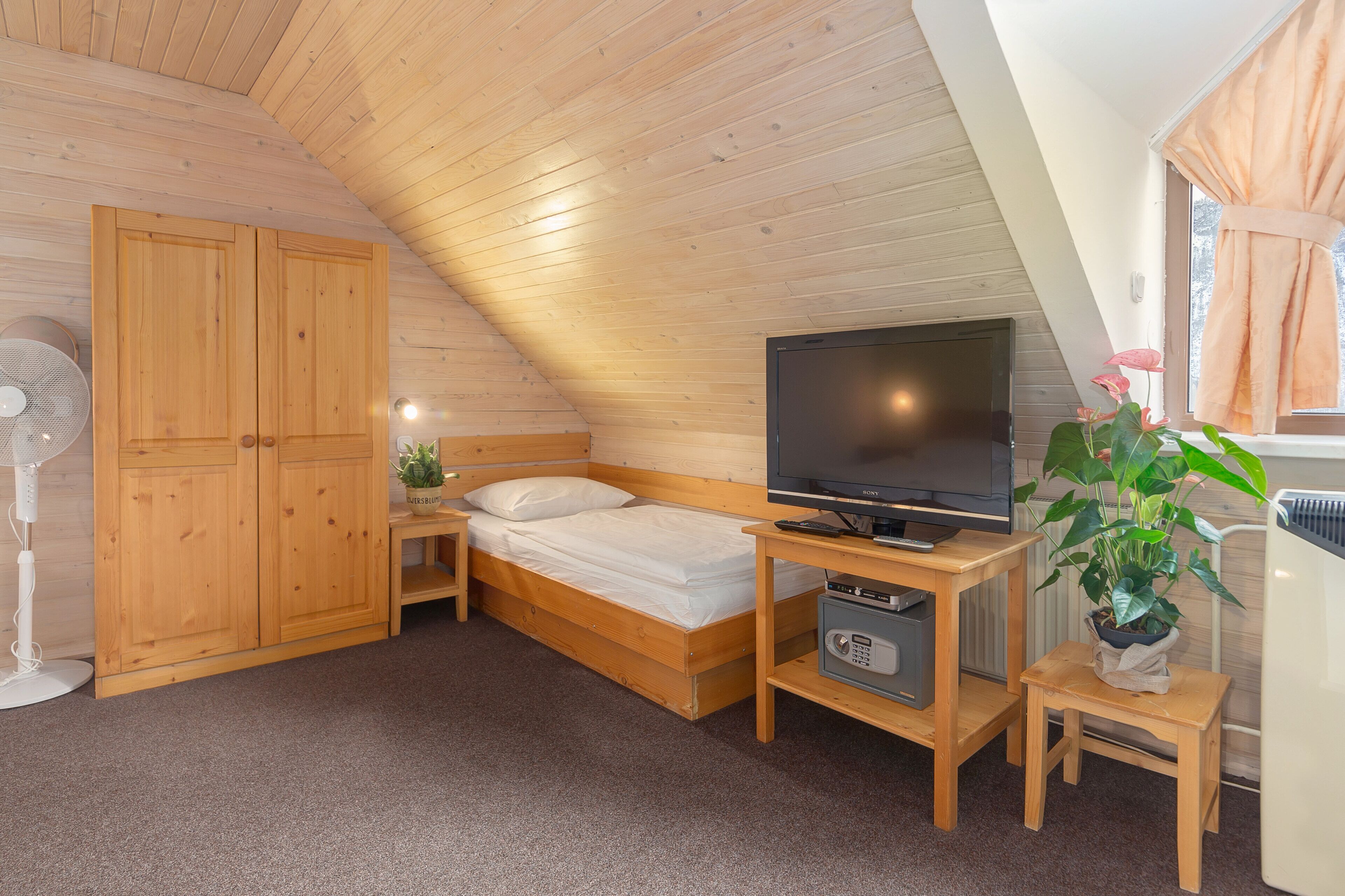 A cozy bedroom with a wooden ceiling, a bed, a TV, a fan, and a potted plant.