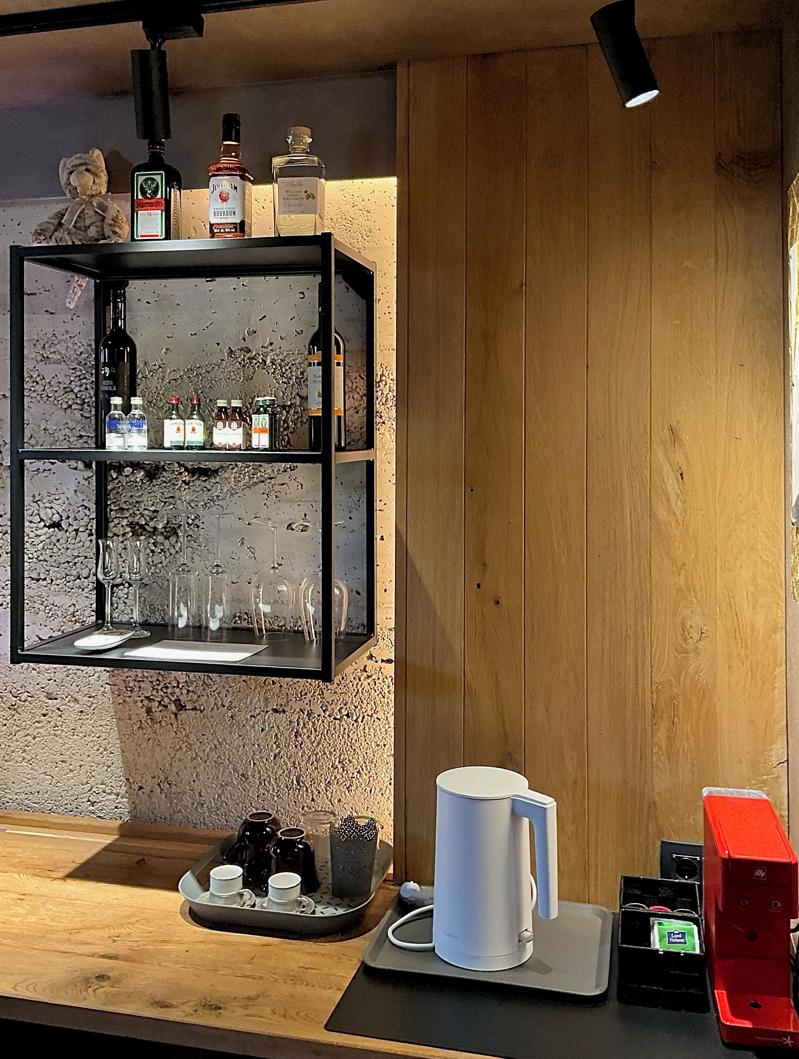 A kitchen area with a wooden counter, a coffee machine, a tray with cups and a teapot, and shelves with bottles and glasses.