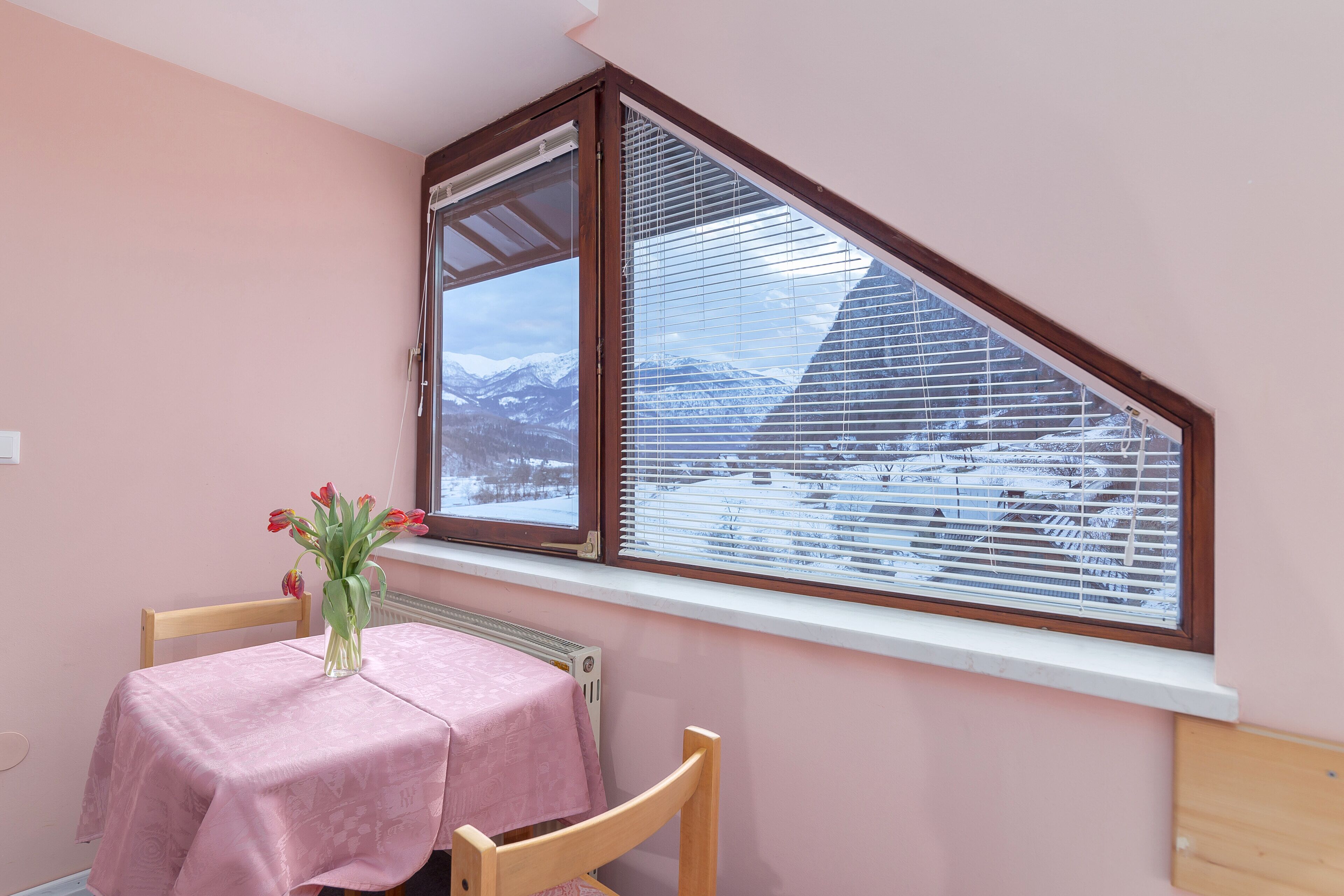 Ein Zimmer mit rosa Wand, einem Fenster mit Jalousien, einem Heizkörper, einem Tisch mit rosa Tischdecke und einem Stuhl.