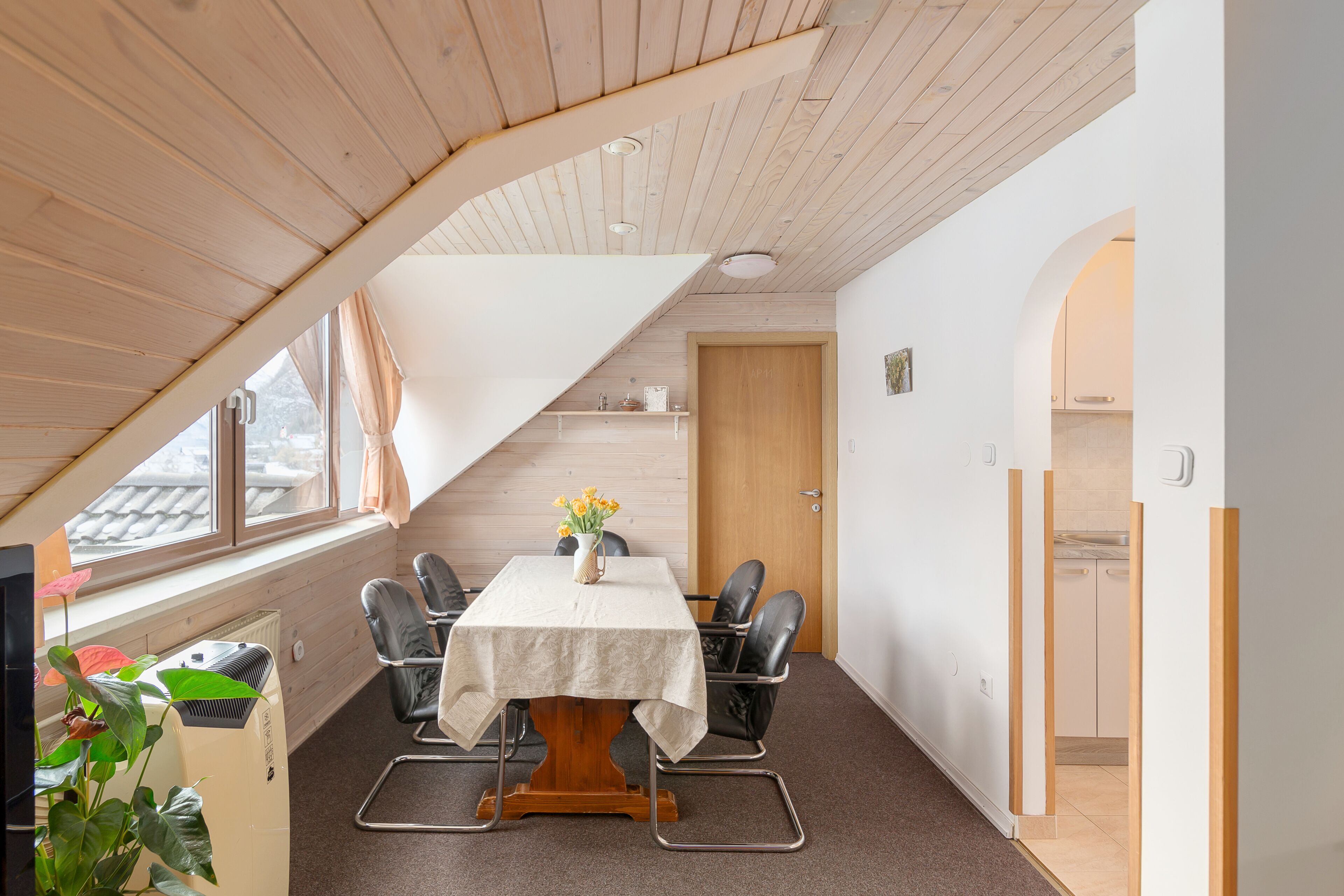 A room with a sloped ceiling, a table with chairs, a potted plant, and a door leading to another room.
