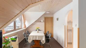 A room with a sloped ceiling, a table with chairs, a potted plant, and a door leading to another room.