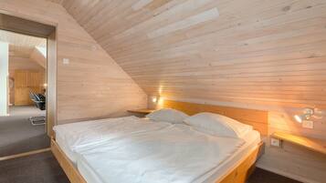 A bedroom with a wooden sloped ceiling, a bed with white bedding, a nightstand with a lamp, and a small wooden shelf.