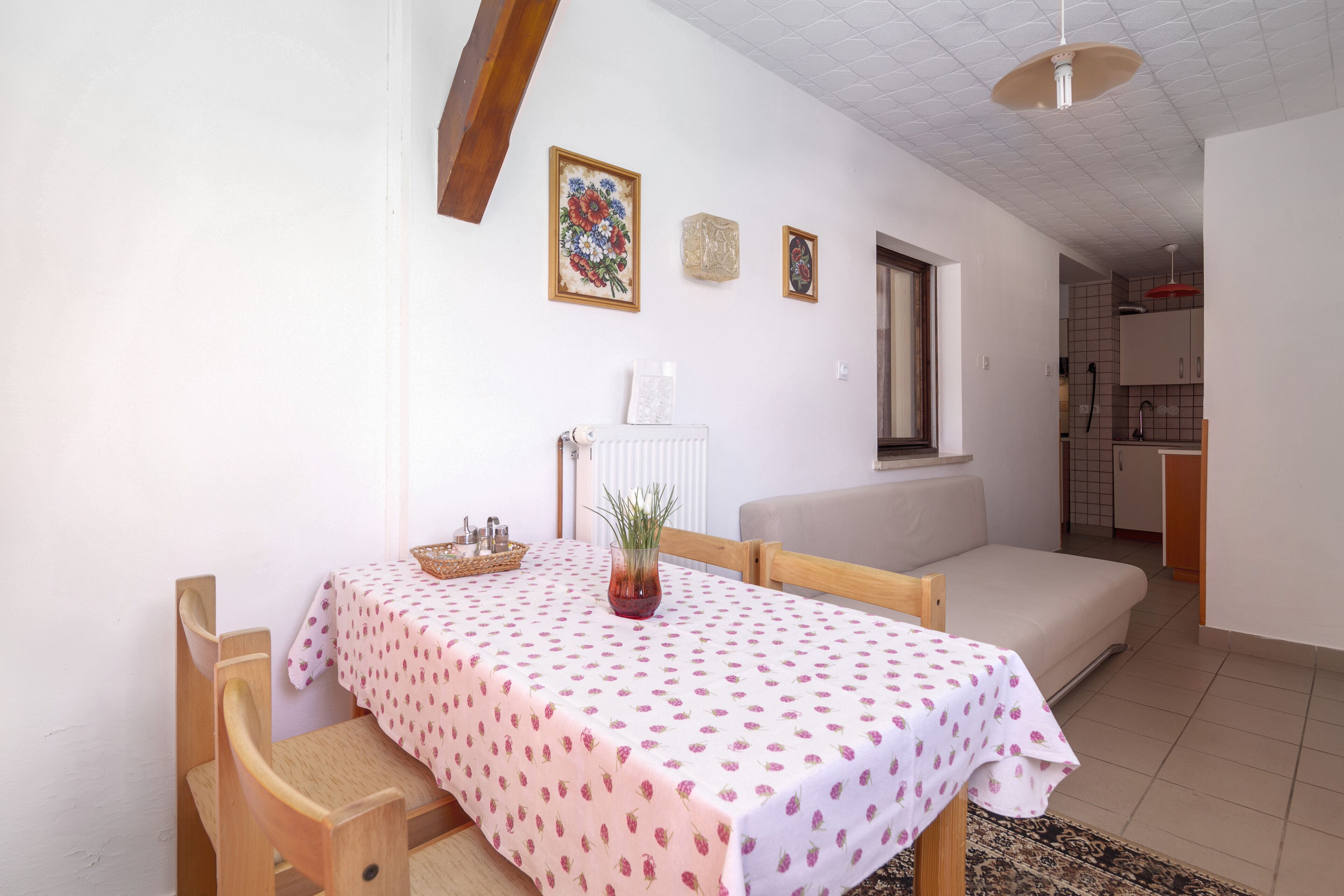 A dining area with a table covered in a polka dot tablecloth, wooden chairs, a vase with a plant, and a radiator.
