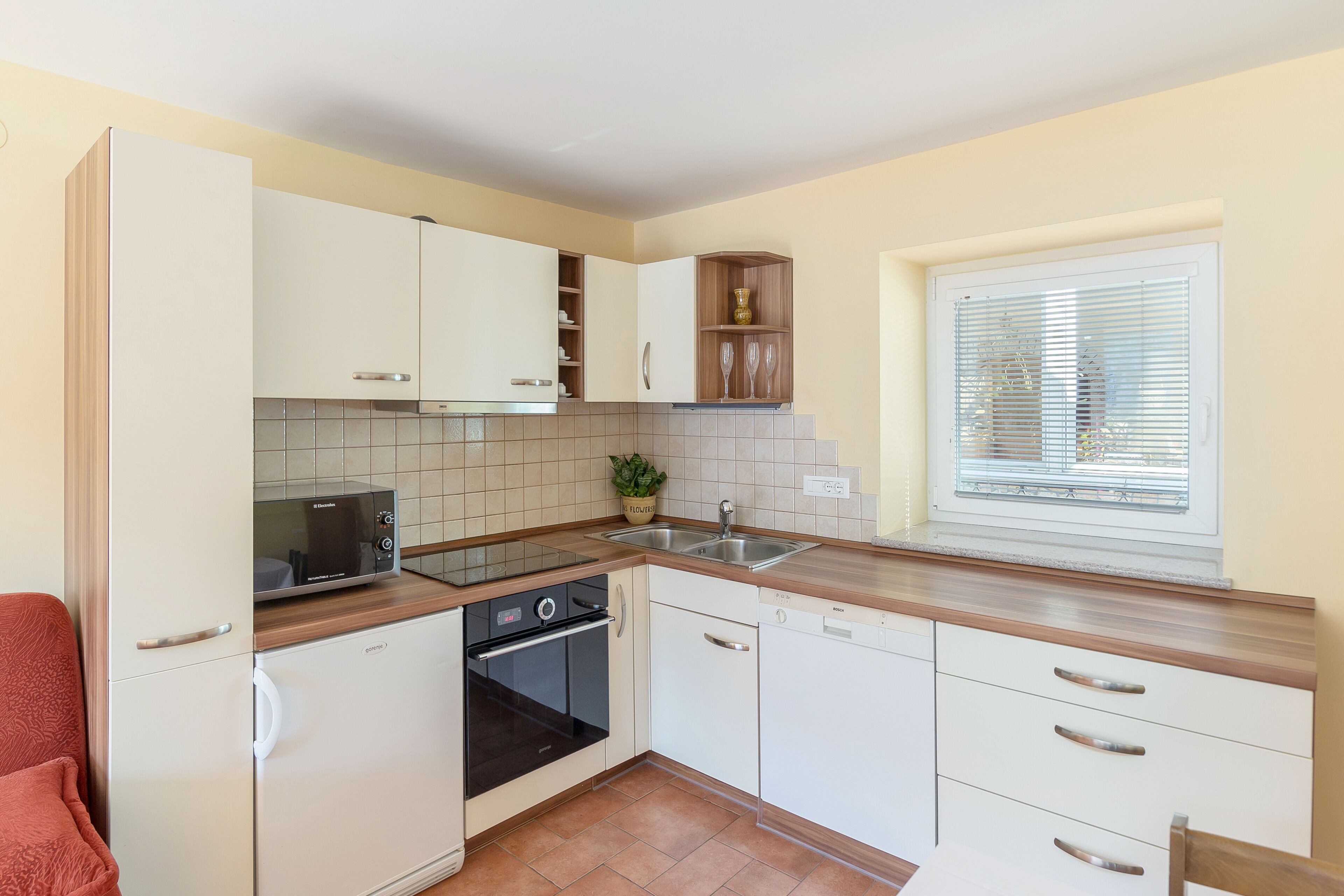 Una cocina compacta con armarios blancos, horno integrado, microondas, lavamanos y una ventana con persianas.
