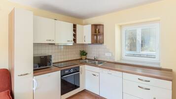 Una cocina compacta con armarios blancos, horno integrado, microondas, lavamanos y una ventana con persianas.