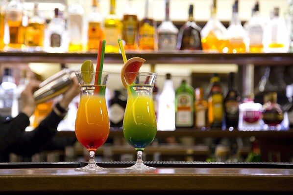 Dos coloridos cócteles sobre una barra, con un fondo desenfocado de varias botellas.