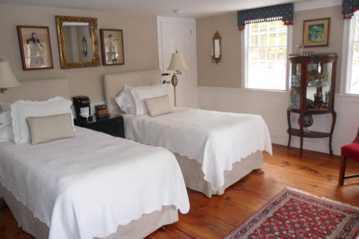 Quarto de hotel com duas camas, piso de madeira, tapete vermelho estampado e mesa de cabeceira de madeira com abajur.