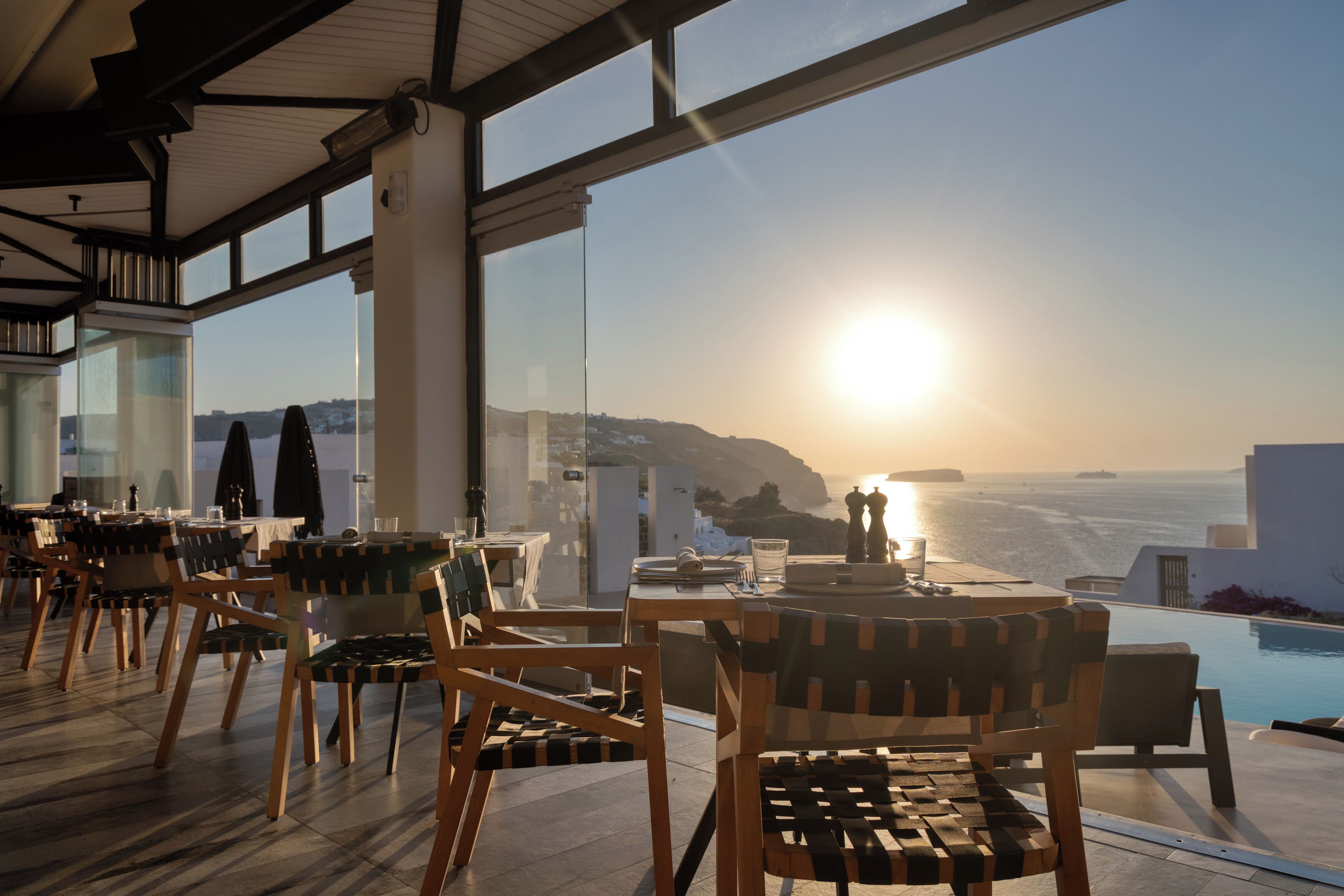 A restaurant with outdoor seating overlooking a pool and the sea at sunset.