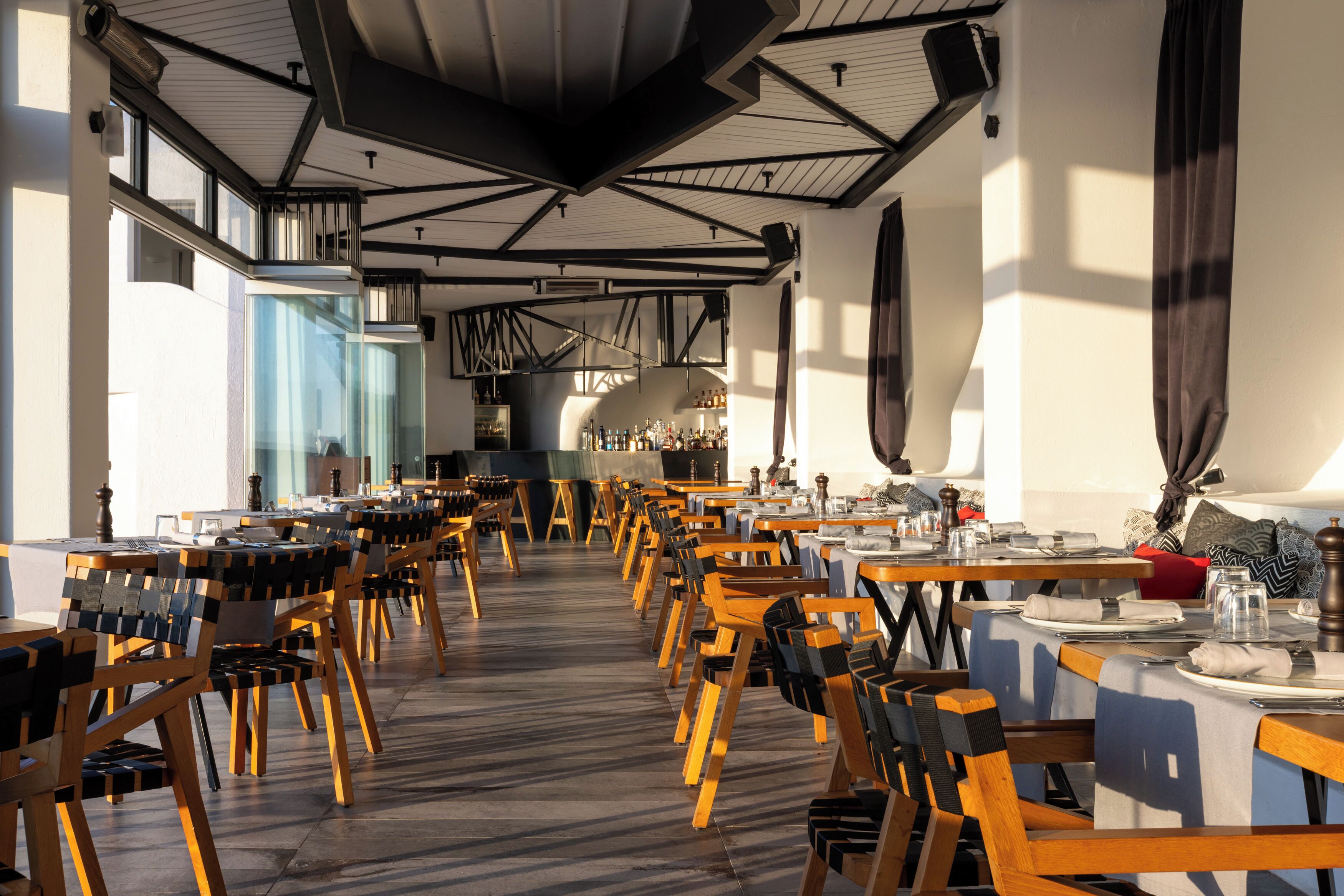 Un espace de restauration intérieur avec plusieurs tables et chaises, un bar avec des bouteilles exposées, et une vue sur la ville grâce à de grandes fenêtres.