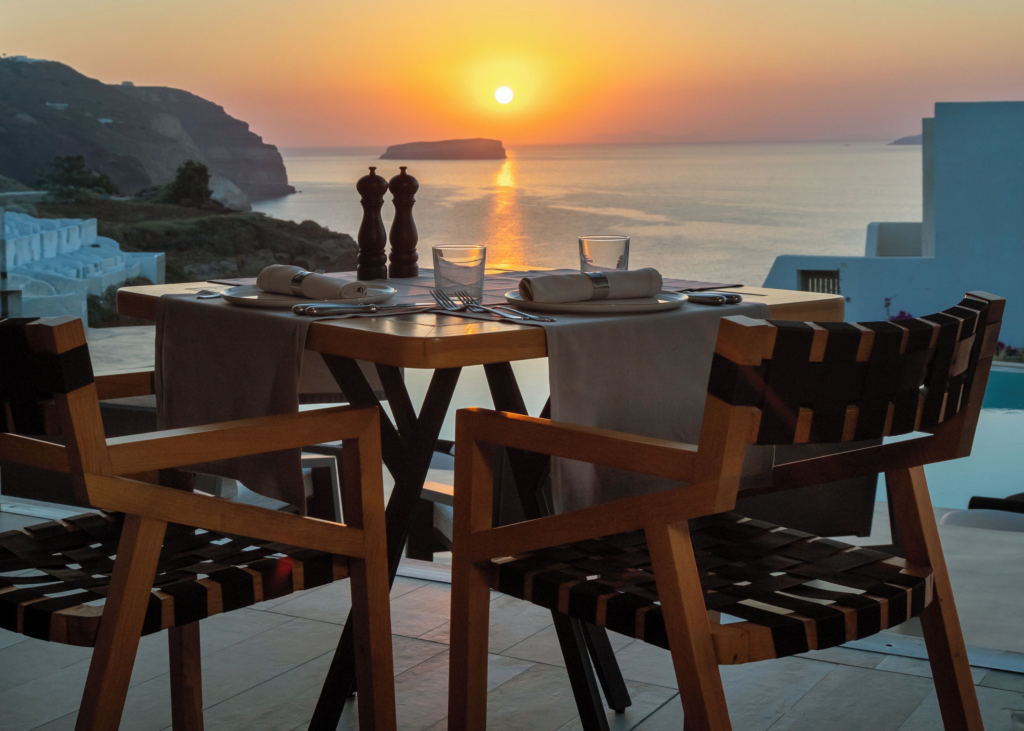 An outdoor dining table with chairs, set against a backdrop of a sunset over the sea.