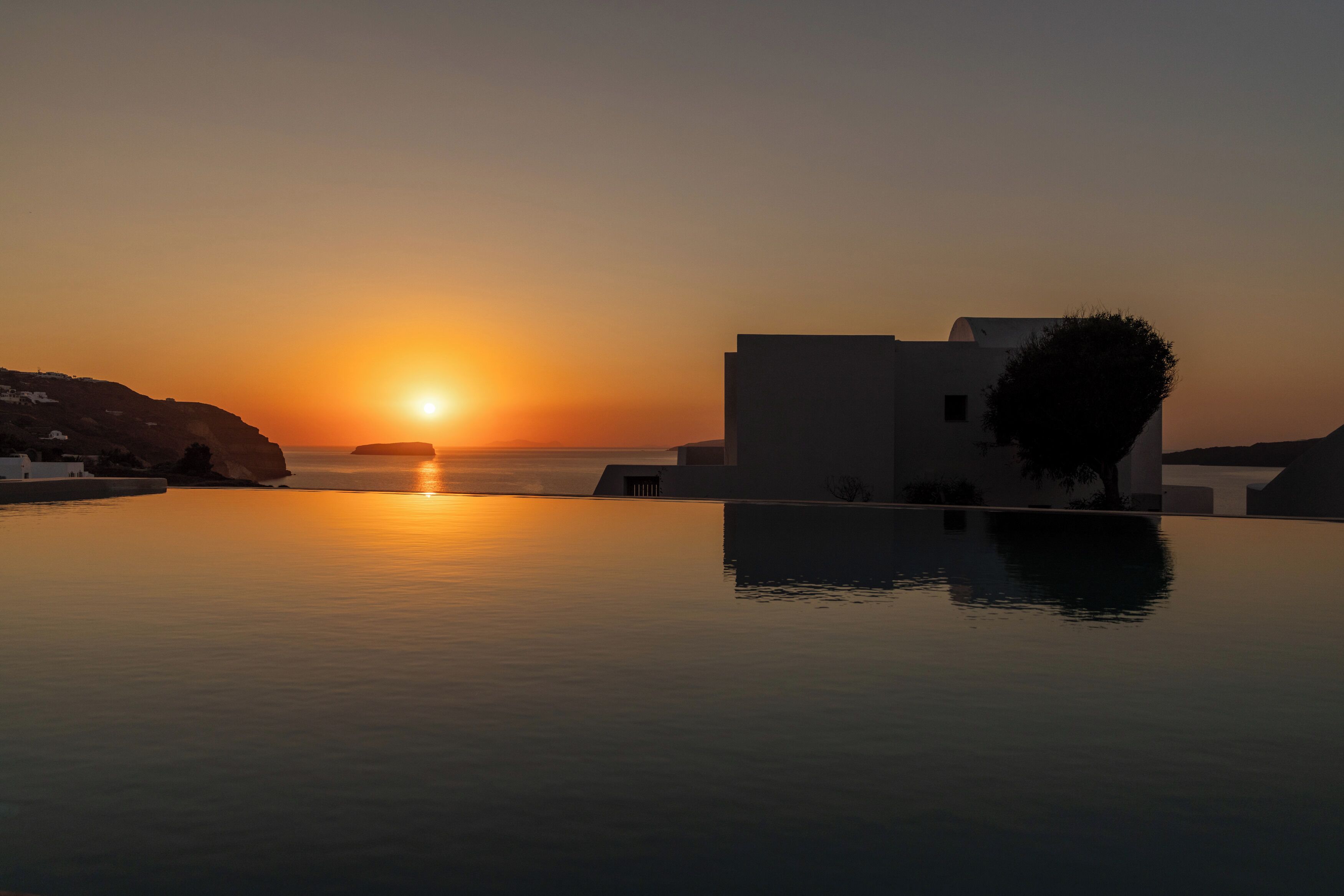 A serene sunset over a calm pool with a building and rocky landscape in the background.