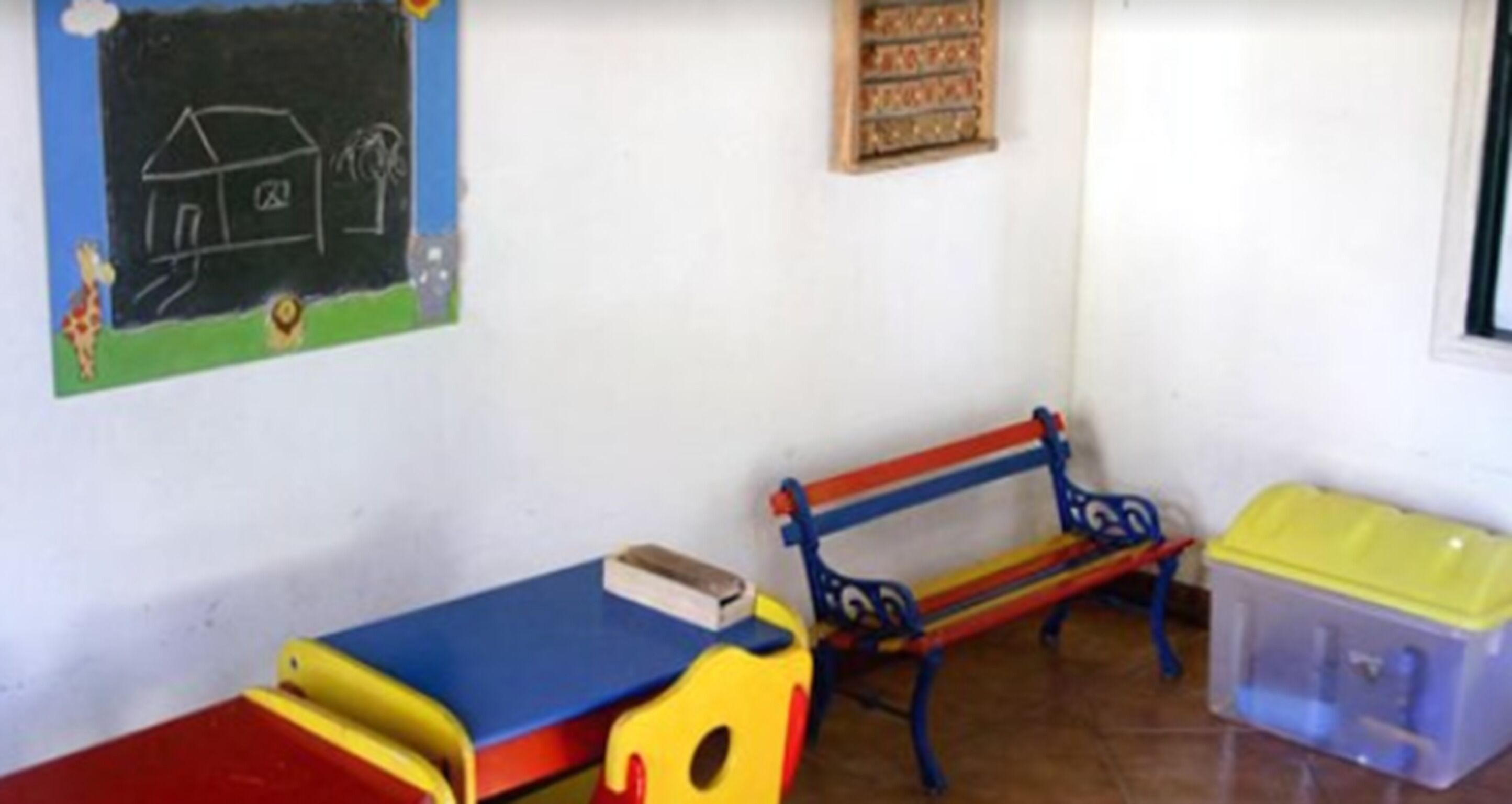 Une pièce avec un banc bleu et rouge, une petite table sur laquelle se trouve un livre, et un récipient en plastique posé sur le sol.