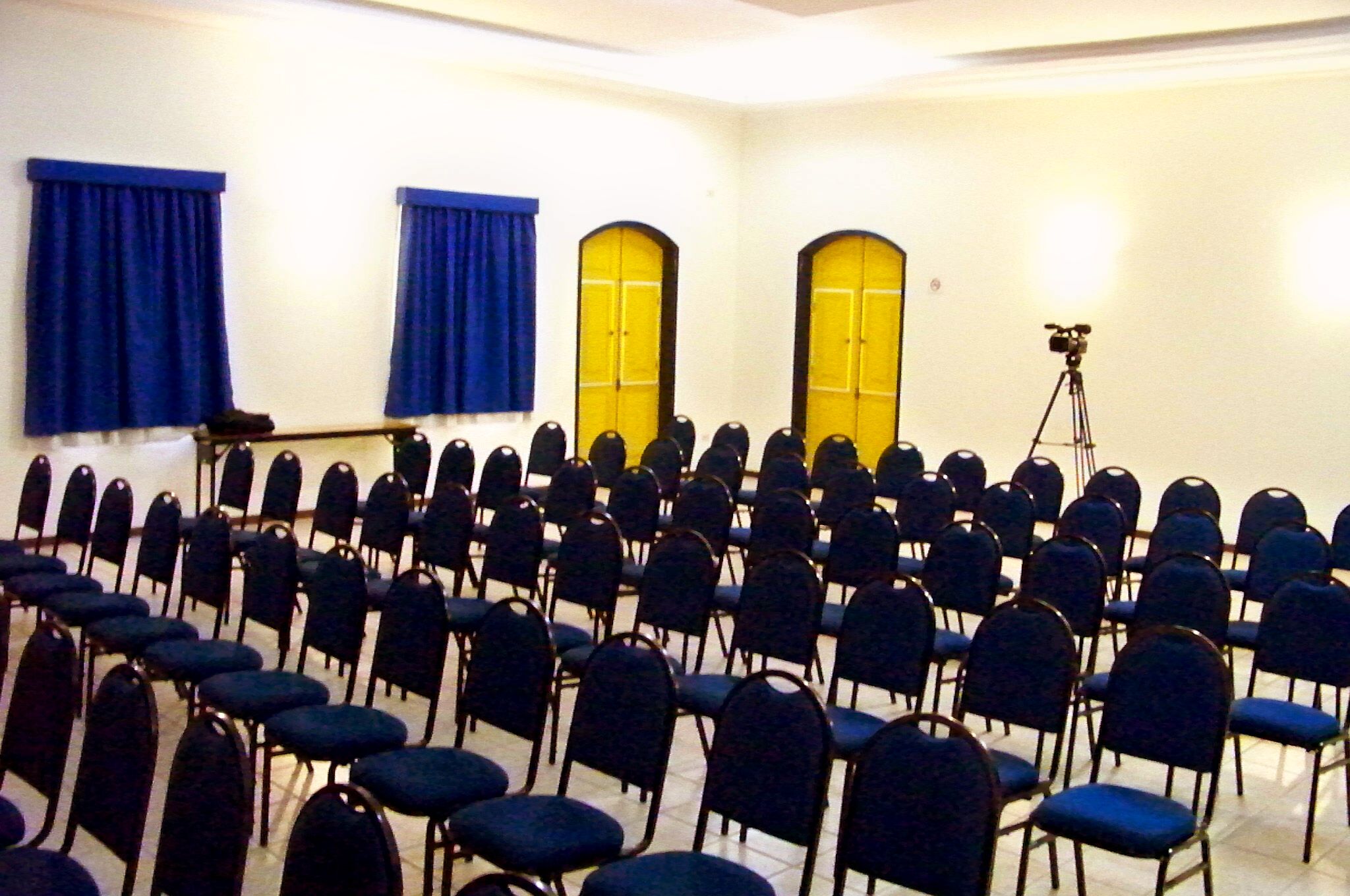 Une salle de conférence avec des rangées de chaises, des rideaux bleus et des portes jaunes.