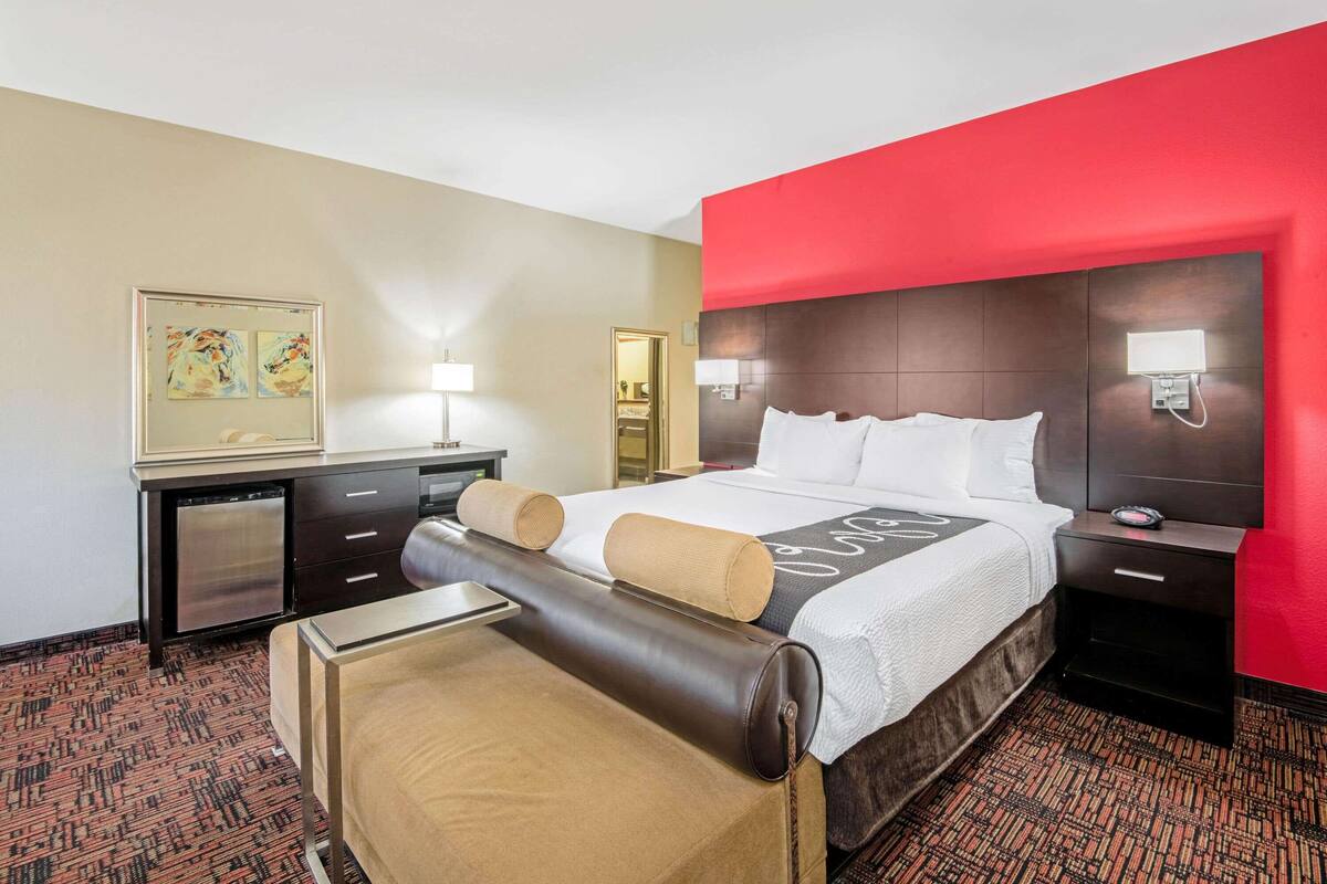 A hotel room with a large bed, a desk with a chair, a desk lamp, and a nightstand with a lamp.