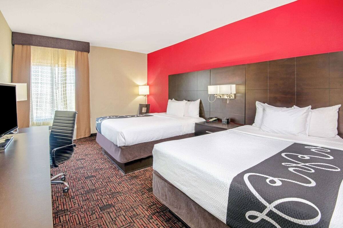 A hotel room with two beds, a desk, a chair, a TV, and a red accent wall.