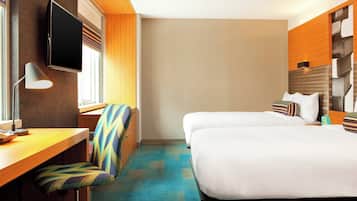 A hotel room with two beds, a desk with a chair, a TV mounted on the wall, and a window with curtains.