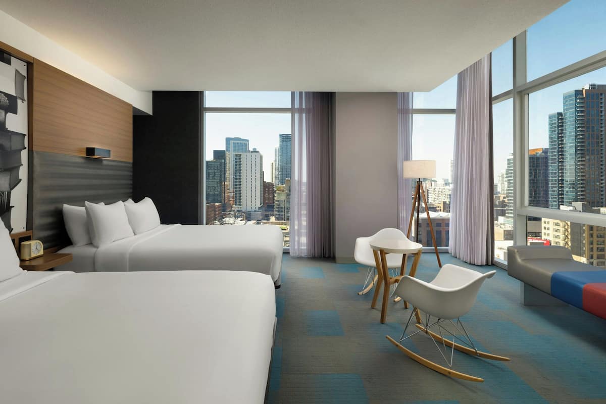 A hotel room with two beds, a desk, a chair, and a view of the cityscape.