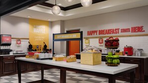 Un espace pour le petit-déjeuner comprenant un comptoir, un coin où l’on prépare des gaufres, et une pancarte indiquant : « ICI COMMENCE UN PETIT-DÉJEUNER JOYEUX ».